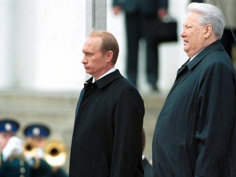 Wladimir Putin und Boris Jelzin stehen nebeneinander auf dem Kathedralenplatz im Kreml. Beide tragen dunkle Anzüge und blicken geradeaus. Die Szene zeigt die feierliche Amtsübergabe am 7. Mai 2000 – Jelzin übergibt das Präsidentenamt an Putin.