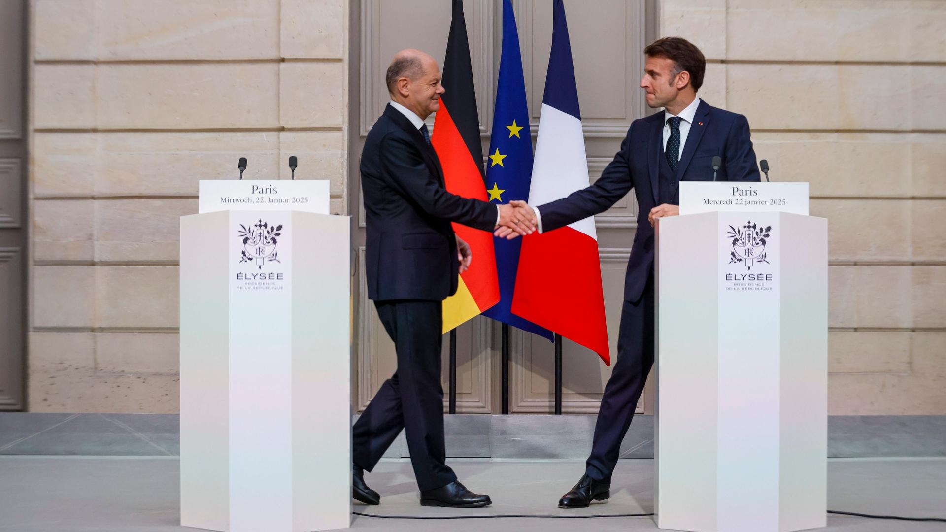 Olaf Scholz und Emmanuel Macron reichen sich die Hand. Sie stehen hinter Rednerpulten und vor einer Türe, vor der die Fahnen der Länder Frankreich und Deutschland hängen sowie die der EU.