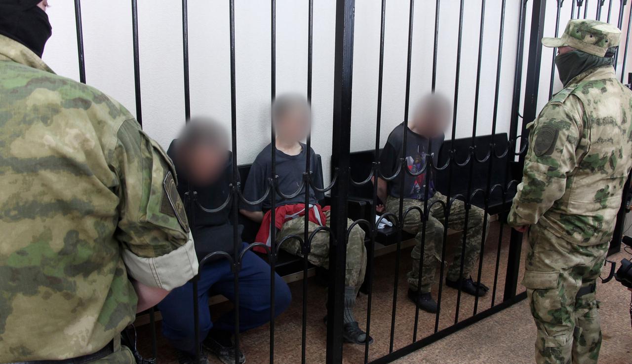 Zwei britische Staatsbürger und ein marokkanischer Staatsbürger sitzen hinter Gittern in einem Gerichtssaal in Donezk. 