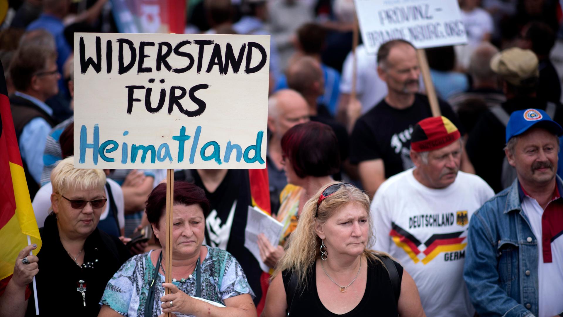Eine Demonstration in Deutschland. Mehrere Menschen stehen zusammen. Eine ältere Frau hält ein Schild in die Höhe. Darauf steht "Widerstand fürs Heimatland". Eine Demonstration in Deutschland. Mehrere Menschen stehen zusammen. Eine ältere Frau hält ein Schild in die Höhe. Darauf steht "Widerstand fürs Heimatland".