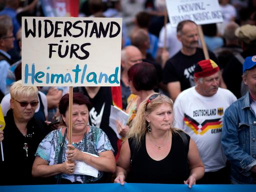 Eine Demonstration in Deutschland. Mehrere Menschen stehen zusammen. Eine ältere Frau hält ein Schild in die Höhe. Darauf steht "Widerstand fürs Heimatland". Eine Demonstration in Deutschland. Mehrere Menschen stehen zusammen. Eine ältere Frau hält ein Schild in die Höhe. Darauf steht "Widerstand fürs Heimatland".