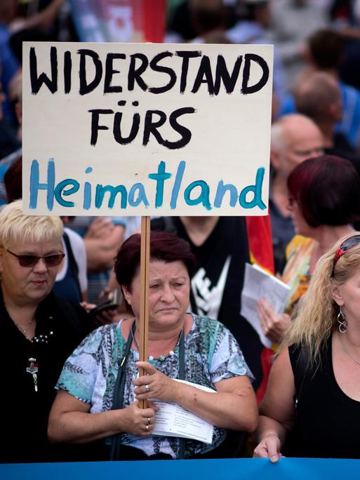 Eine Demonstration in Deutschland. Mehrere Menschen stehen zusammen. Eine ältere Frau hält ein Schild in die Höhe. Darauf steht "Widerstand fürs Heimatland". Eine Demonstration in Deutschland. Mehrere Menschen stehen zusammen. Eine ältere Frau hält ein Schild in die Höhe. Darauf steht "Widerstand fürs Heimatland".