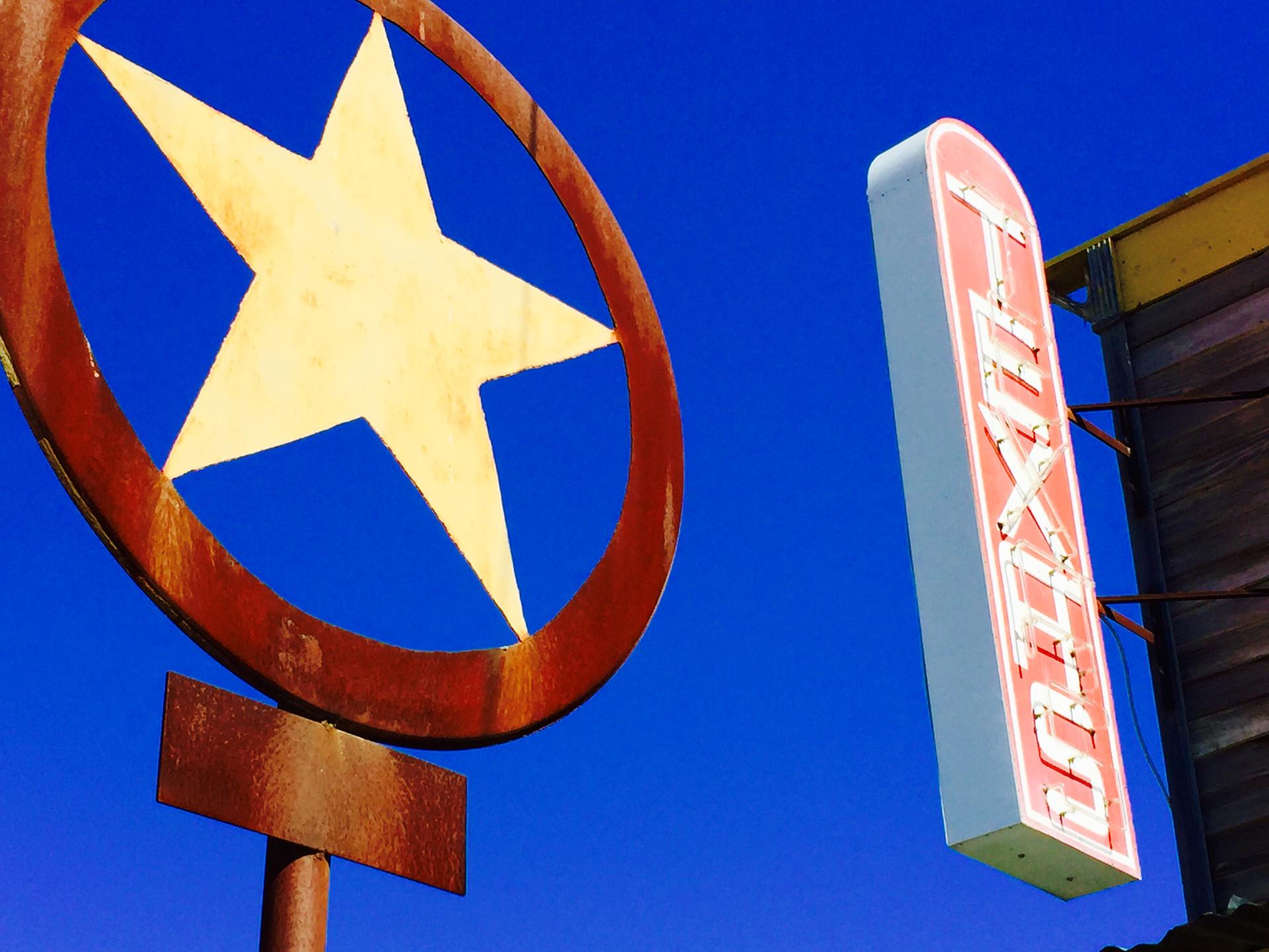 Ein rostiger Texas Lone Star und ein Texas-Schriftzug in Leuchtschrift vor einem intensivblauen Himmel.