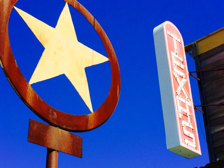 Ein rostiger Texas Lone Star und ein Texas-Schriftzug in Leuchtschrift vor einem intensivblauen Himmel.