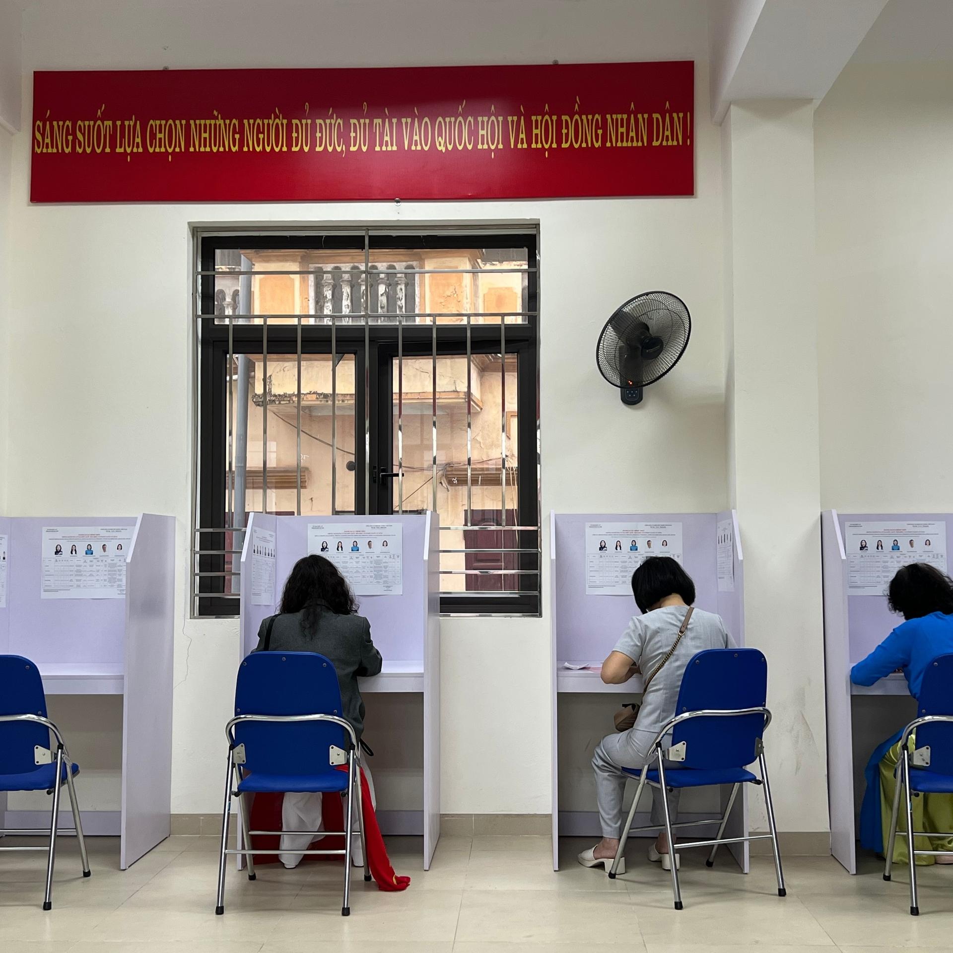 Menschen sitzen in Wahlkabinen, die vor einer Wand aufgereiht sind. 