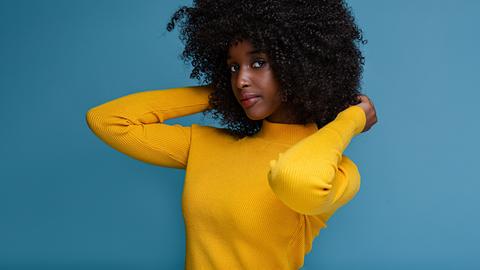 Eine weibliche Person of Color steht in gelbem Pullver vor einer blauen Wand.