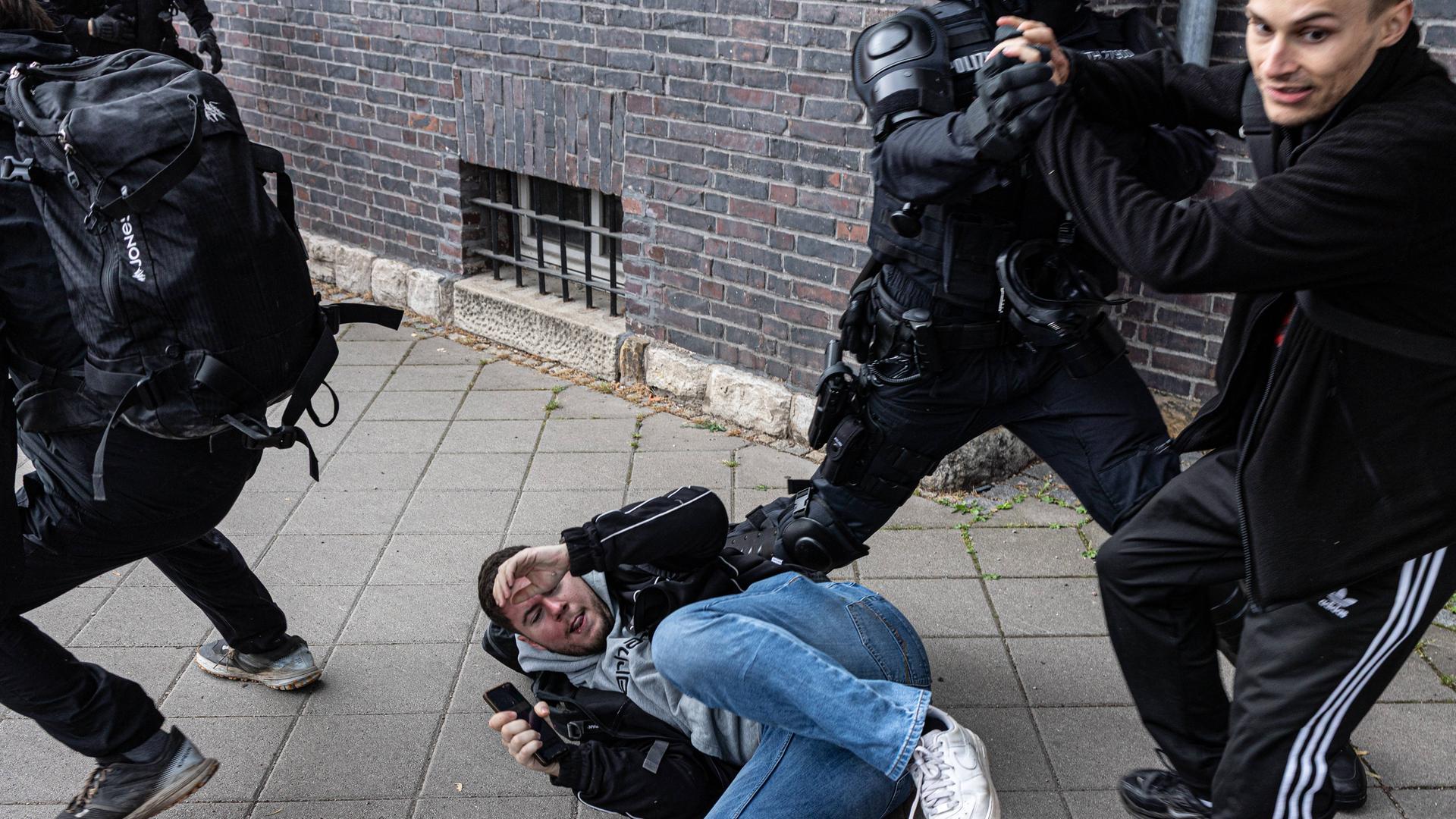 Ein Vertreter von Apollo News wird bei den Protesten gegen den AfD-Parteitag am Wochenende angegriffen. Polizisten versuchen, ihn zu schützen. Das Bild zeigt einen Mann, am Boden liegend und einen weiteren, der von einem Polizisten zurückgedrängt wird.