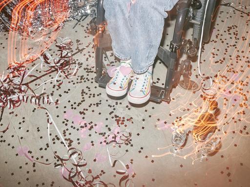 Nahaufnahme der Beine einer Frau in Turnschuhen, die in einem Rollstuhl sitzt, umgeben von Konfetti, Luftschlangen und leeren Gläsern auf einer Tanzfläche.