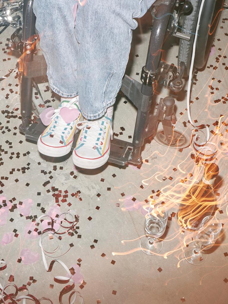 Nahaufnahme der Beine einer Frau in Turnschuhen, die in einem Rollstuhl sitzt, umgeben von Konfetti, Luftschlangen und leeren Gläsern auf einer Tanzfläche.