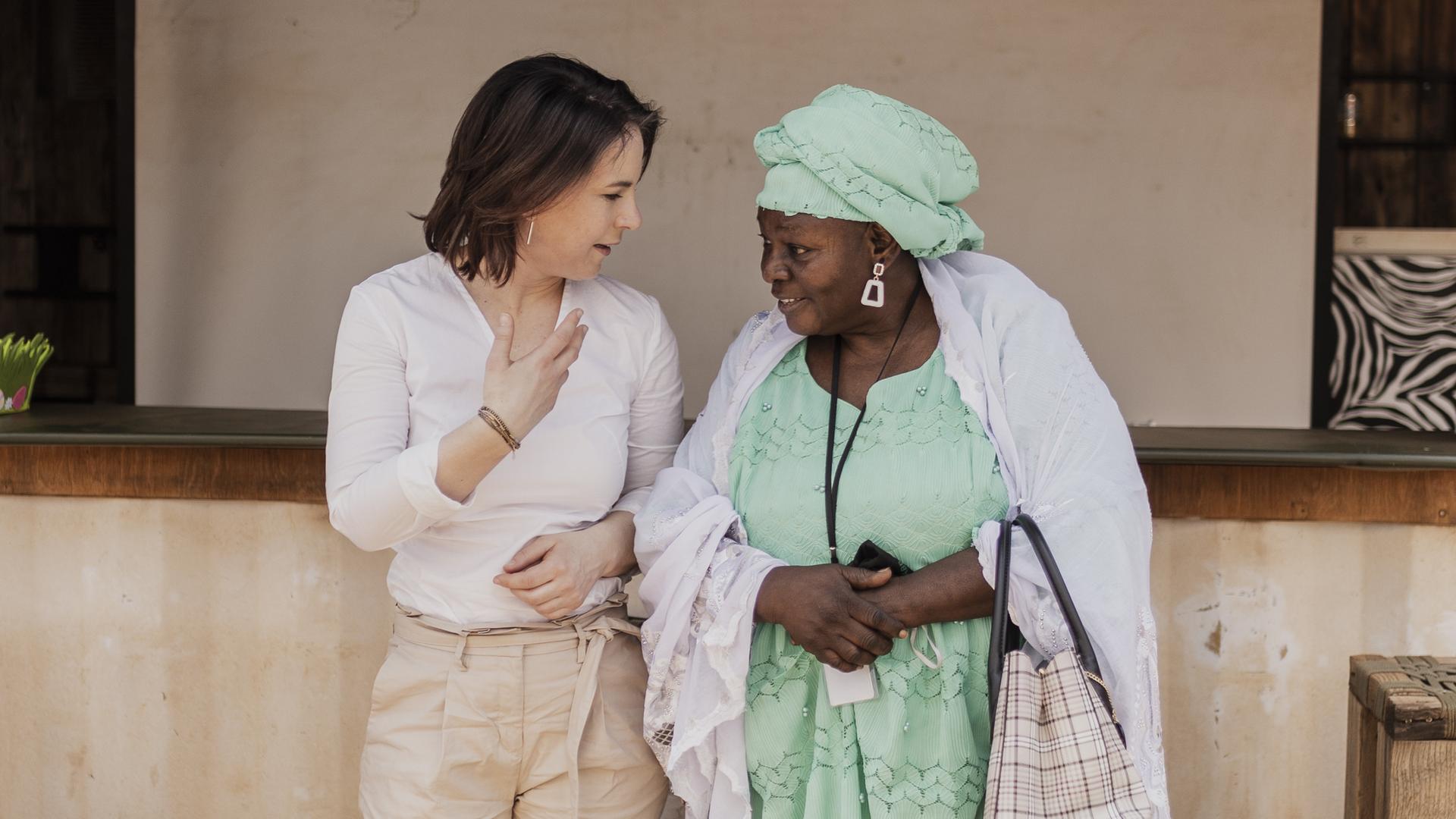 Die Bundesaussenministerin Annalena Bearbock steht mit einer Frau in traditioneller Tracht vor einer Hauswand, Mali 2022. 
