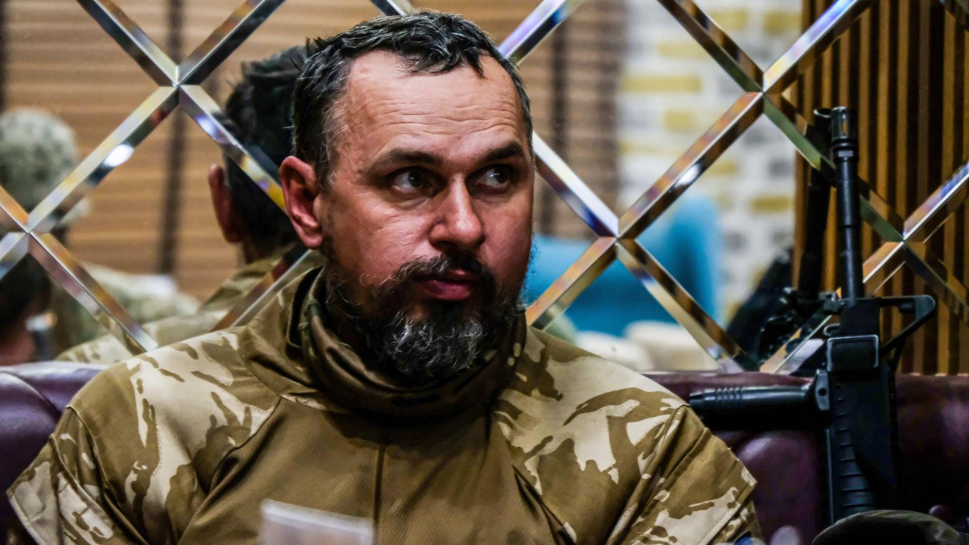 Der ukrainische Filmregisseur Senzow sitzt in einer Militäruniform vor einem Gittertor. Links neben ihm steht ein Gewehr. Der ukrainische Filmregisseur Senzow sitzt in einer Militäruniform vor einem Gittertor. Links neben ihm steht ein Gewehr.