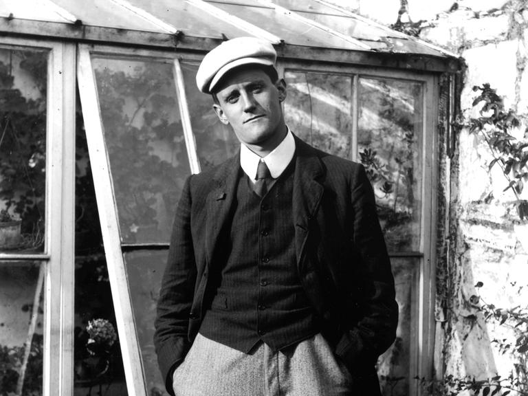 Der Schriftsteller James Joyce steht vor einem Gewächshaus, trägt Schiebermütze und schaut in die Kamera.
