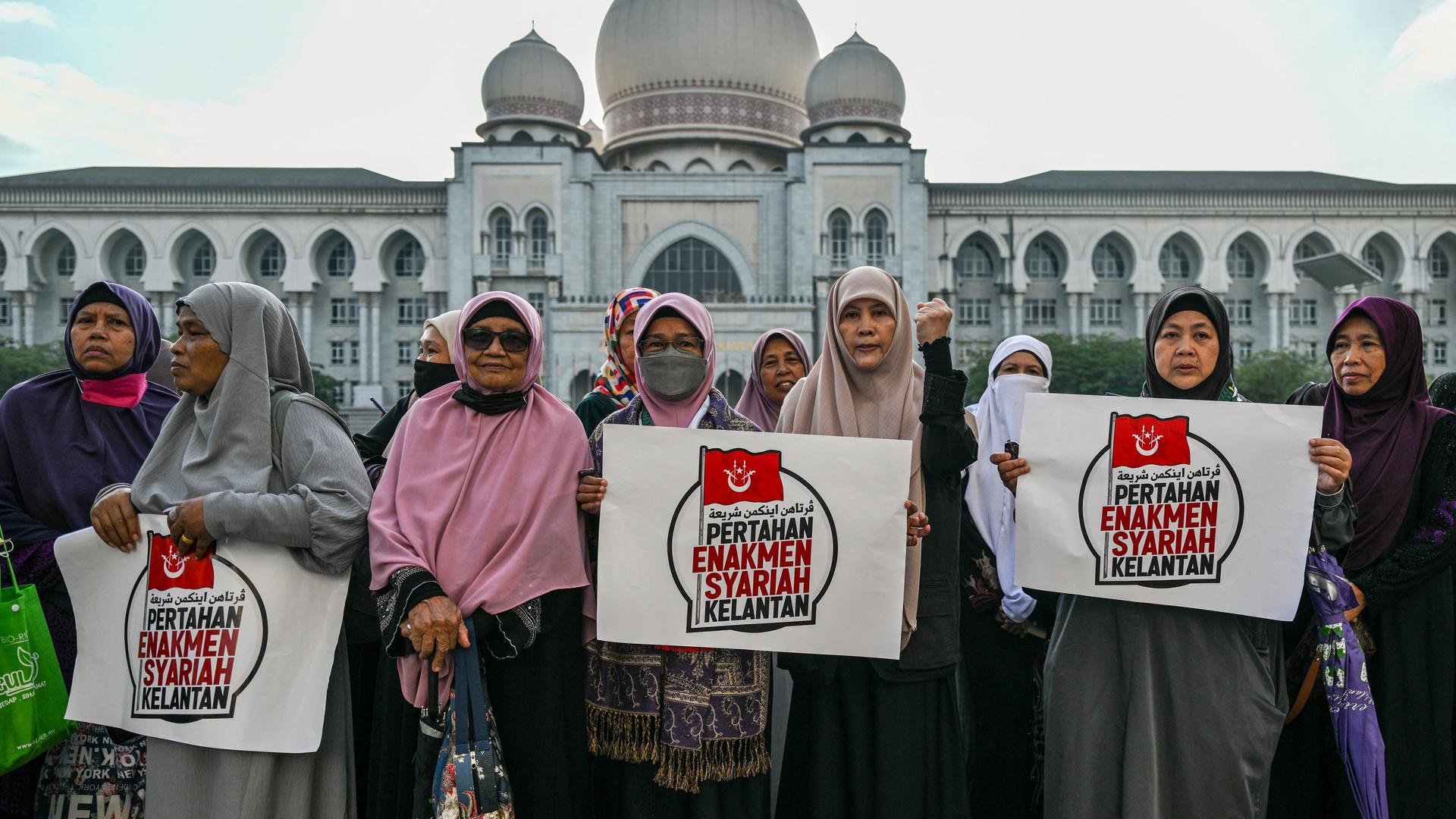 Frauen mit Kopftüchern stehen vor einem Gebäude und halten Protest-Schilder hoch.