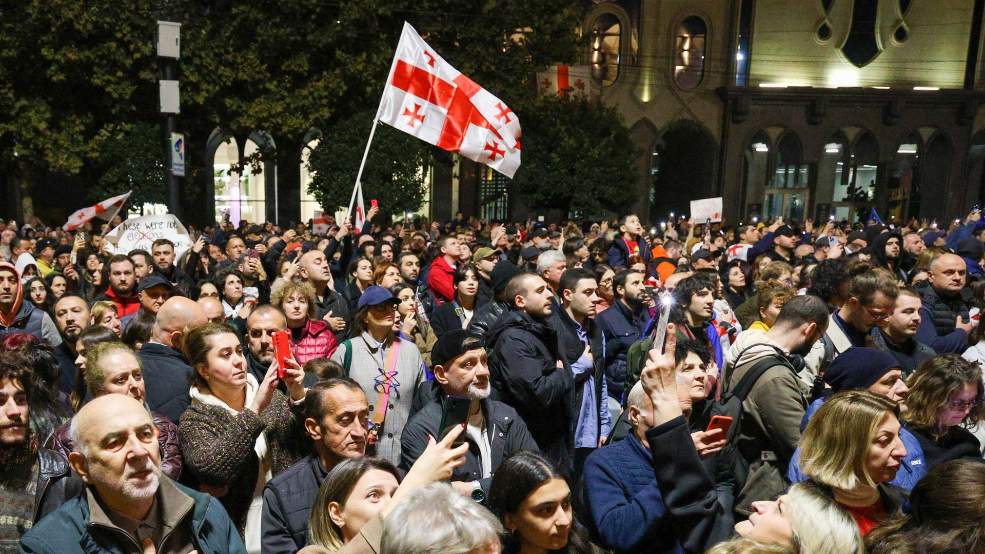 Viele Menschen stehen im Dunkeln auf einer Straße. Manche schwenken die georgische Flagge. 