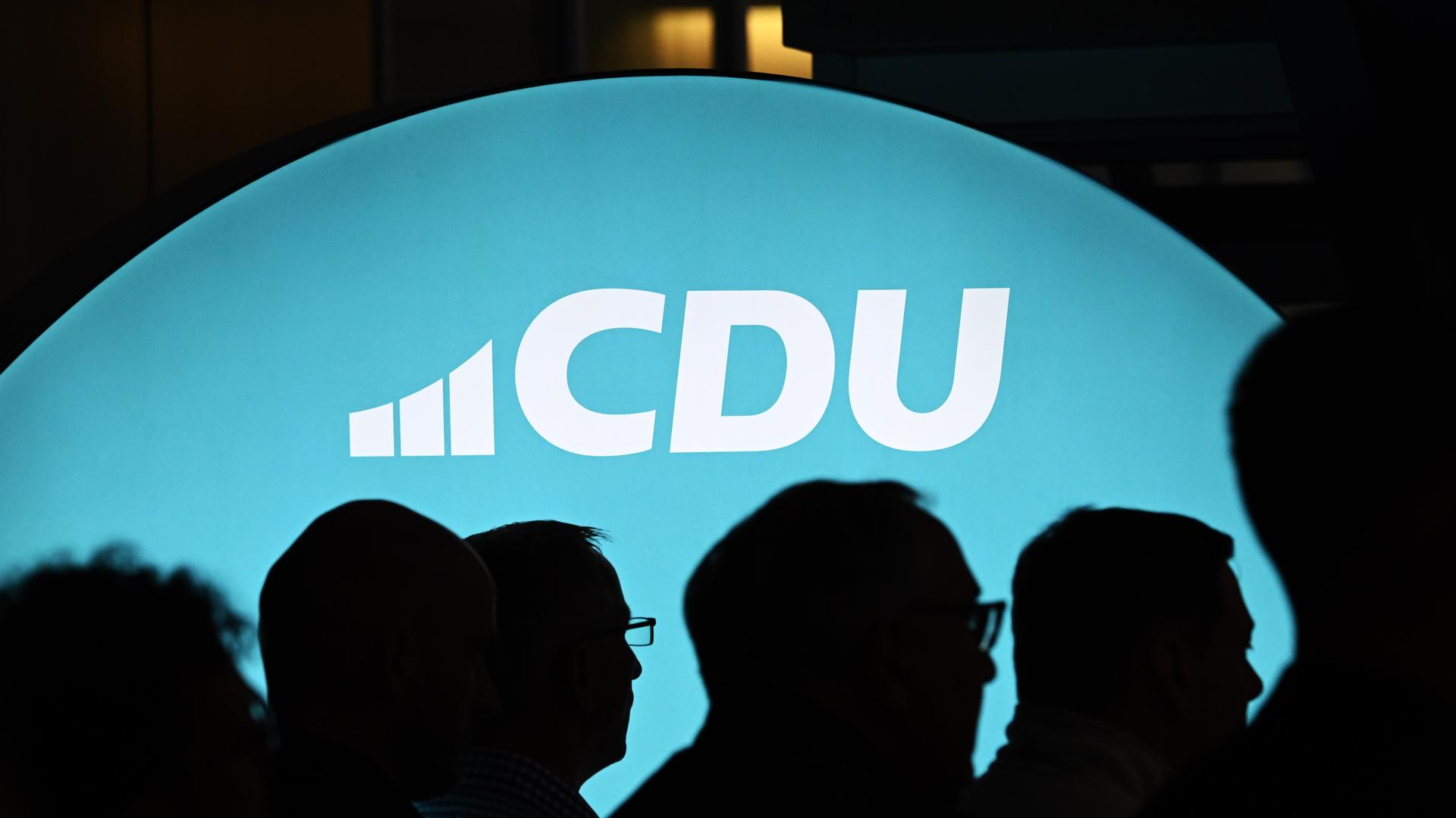 Ein beleuchtetes Banner mit dem Schriftzug "CDU" ist zu sehen, davor sieht man im Schatten die Köpfe mehrerer Menschen.