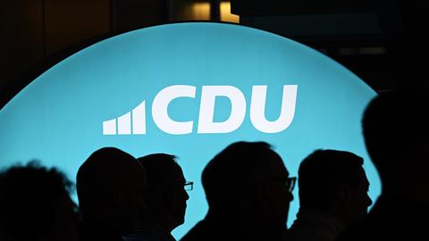 Ein beleuchtetes Banner mit dem Schriftzug "CDU" ist zu sehen, davor sieht man im Schatten die Köpfe mehrerer Menschen.