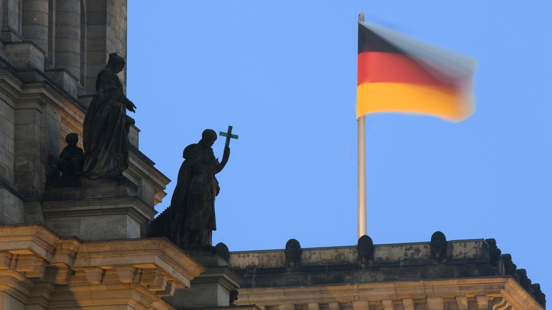 Auf dem Reichstagsgebäude weht eine Deutsche Fahne und eine Statue aus Stein hält ein Kreuz als Symbol für die christliche Kirche, Berlin, Deutschland, Europa Auf dem Reichstagsgebäude weht eine Deutsche Fahne und eine Statue aus Stein hält ein Kreuz als Symbol für die christliche Kirche, Berlin, Deutschland, Europa