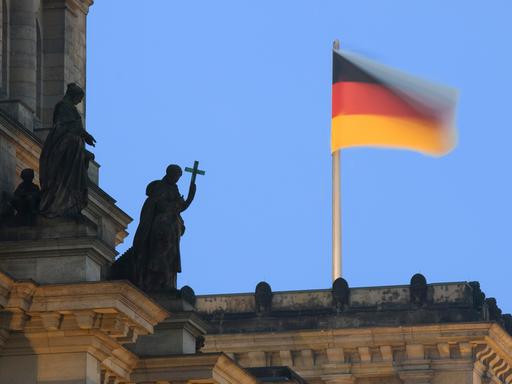 Auf dem Reichstagsgebäude weht eine Deutsche Fahne und eine Statue aus Stein hält ein Kreuz als Symbol für die christliche Kirche, Berlin, Deutschland, Europa