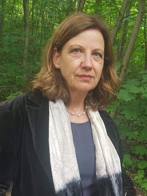 Eine Frau in einem Park, seitlich zu sehen, mit weißem Schal über einem schwarzen Samtjackett. Sie hat braune Haare und blickt nachdenklich.