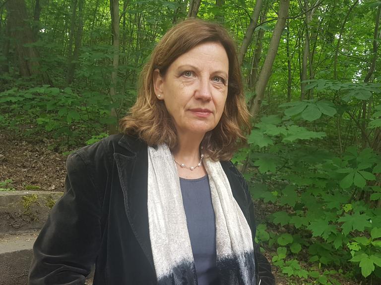 Eine Frau in einem Park, seitlich zu sehen, mit weißem Schal über einem schwarzen Samtjackett. Sie hat braune Haare und blickt nachdenklich.