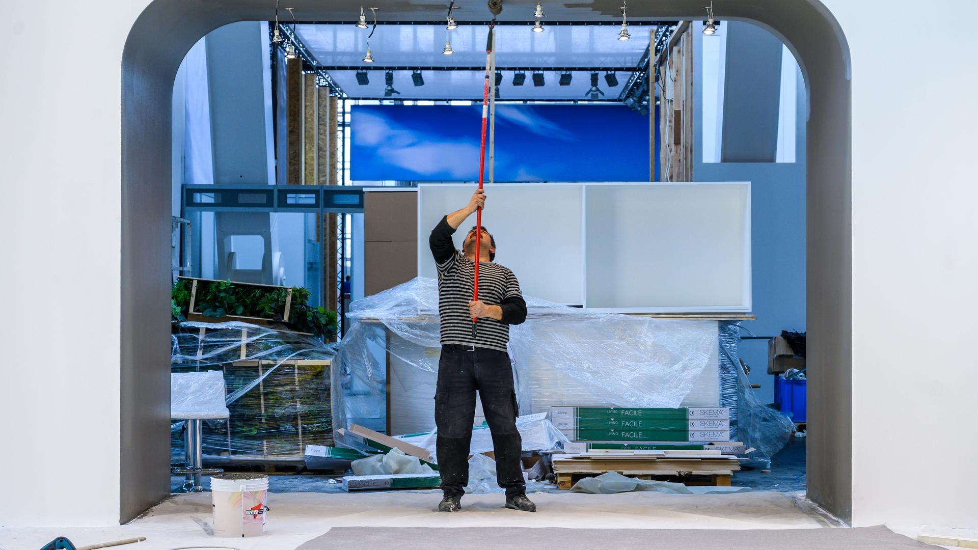 Ein Handwerker steht in einem Durchgang einer im Aufbau befindlichen Messehalle und versucht mit einem langen Besenstiel die Decke zu erreichen.