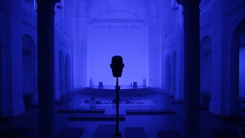 Ein Saal (blau eingetönt) mit einem großen Lautsprecher in der Mitte, rechts und links je eine Säule. 
