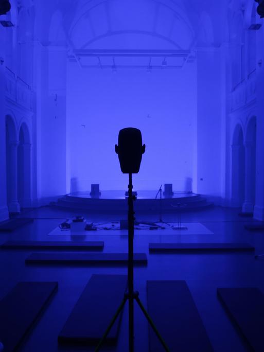 Ein Saal (blau eingetönt) mit einem großen Lautsprecher in der Mitte, rechts und links je eine Säule. 