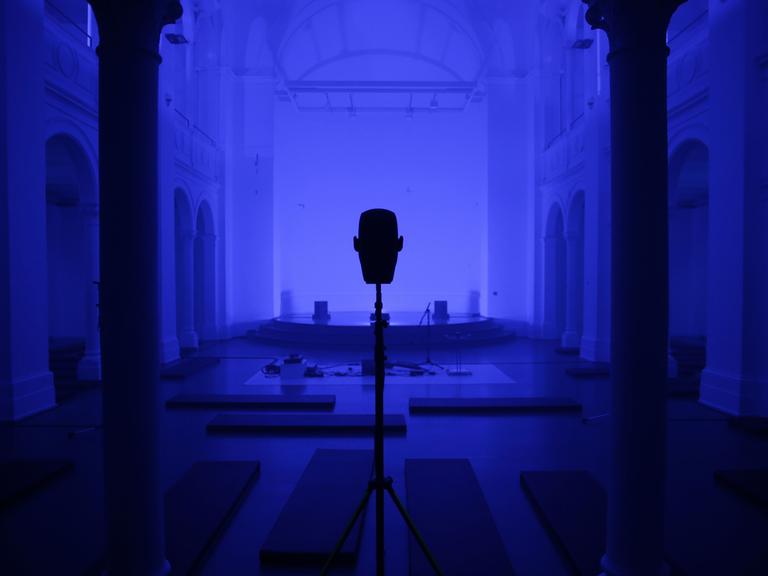 Ein Saal (blau eingetönt) mit einem großen Lautsprecher in der Mitte, rechts und links je eine Säule. 