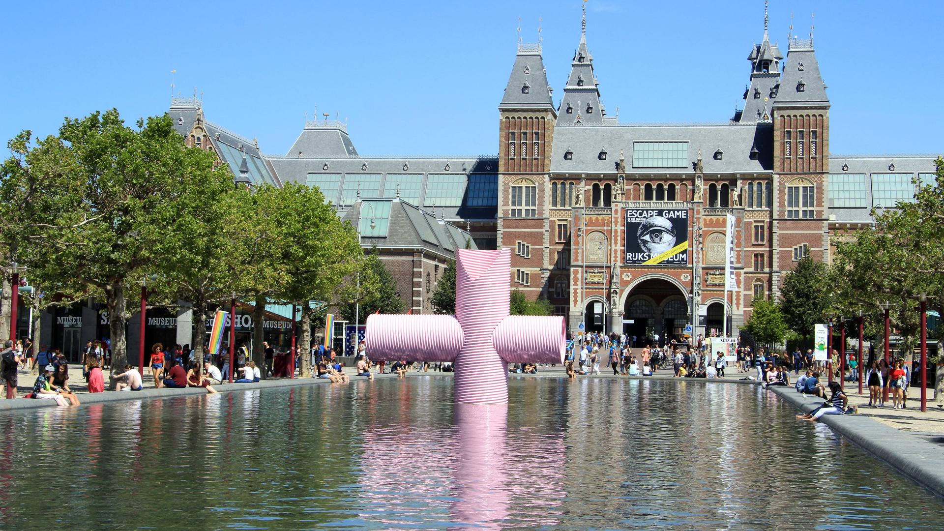 Blick auf das Rijksmuseum in Amsterdam; ein großes altes Gebäude. Im Vordergrund ist ein Wasserbecken mit einer Kunstskulptur.