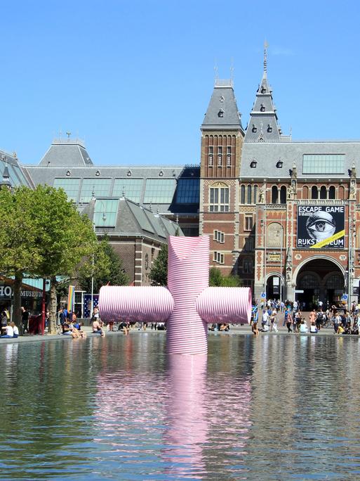 Blick auf das Rijksmuseum in Amsterdam; ein großes altes Gebäude. Im Vordergrund ist ein Wasserbecken mit einer Kunstskulptur.