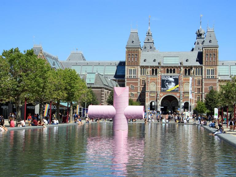 Blick auf das Rijksmuseum in Amsterdam; ein großes altes Gebäude. Im Vordergrund ist ein Wasserbecken mit einer Kunstskulptur.