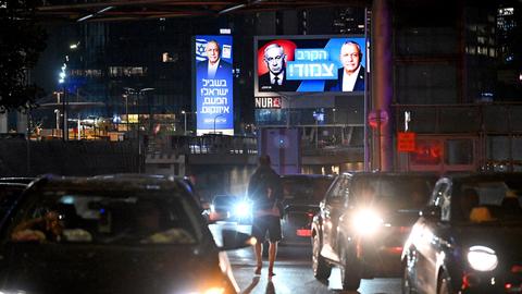 Tel Aviv bei Nacht: Mehrere Autos fahren auf einer Straße, an einem Gebäude im Hintergrund sind zwei erleuchtete Wahlplakate zu sehen.