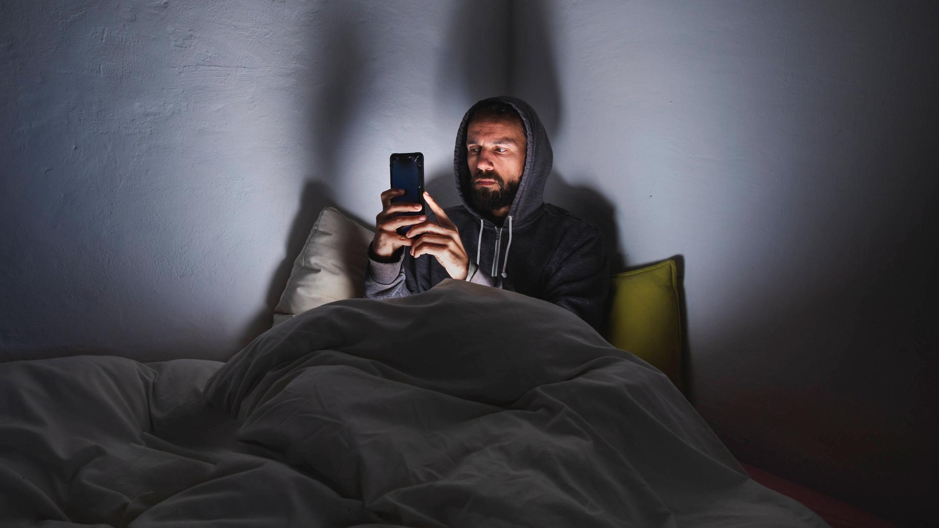 Ein Mann sitzt in einem Bett und schaut auf sein Handy. Im Zimmer ist es dunkel, aber das Handy erhellt den Raum. 