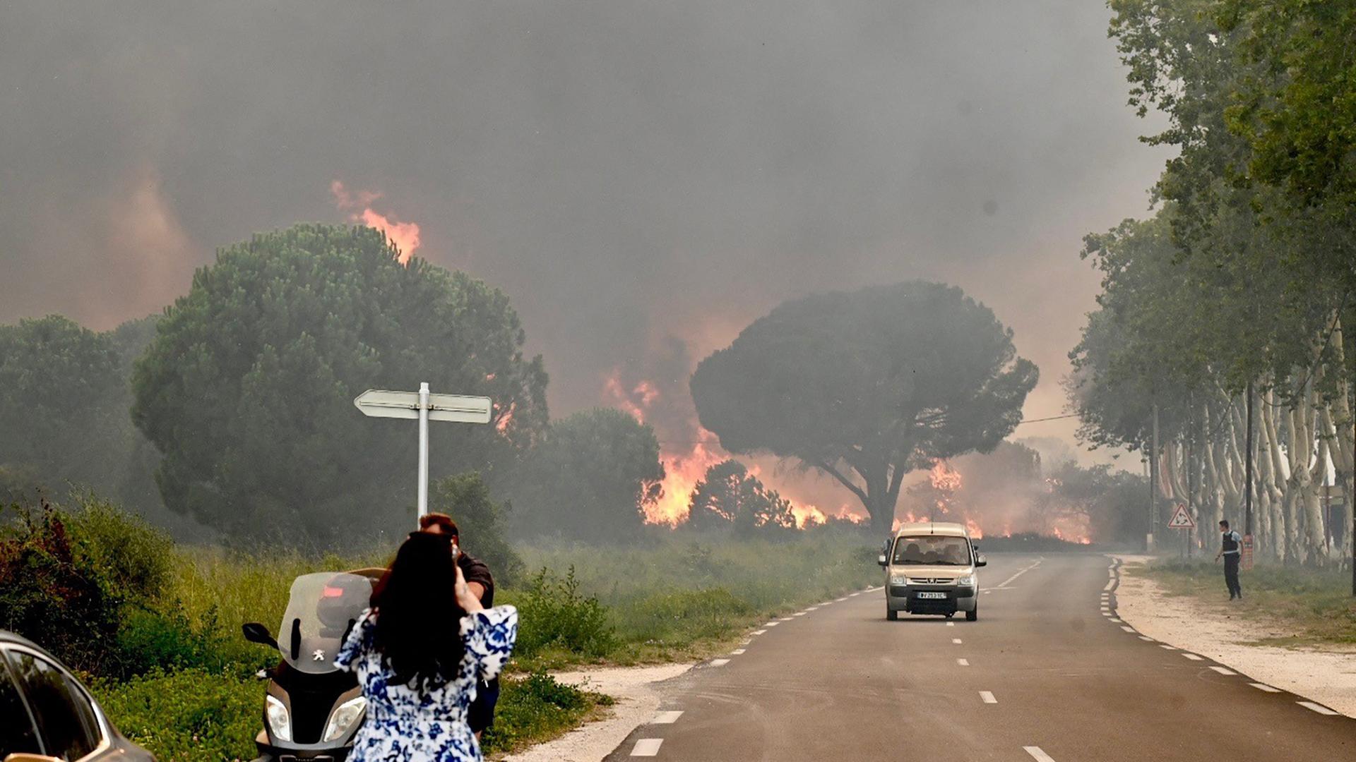 Blick auf ein brennendes Waldstück. Ein Auto fährt in Richtung der Kamera, um aus der Gefahrenzone zu gelangen. Am Straßenrand steht eine Frau, die aus der Entfernung ein Foto von dem Feuer schießt.