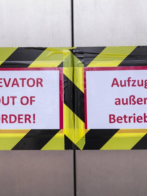 Blick auf die geschlossenen Türen eines Berliner Fahrstuhls, links ein mit Klebeband befestigtes Schild mit der Aufschrift "Elevator out of order!", rechts "Fahrstuhl außer Betrieb!"