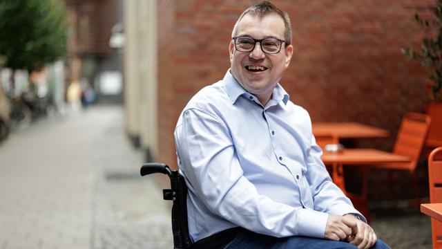 Jürgen Linnemann sitzt in einem Rollstuhl vor einem roten Gebäude. Er lächelt.