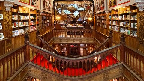Innenansicht einer der schönsten Buchhandlungen der Welt: Die Livraria Lello in Porto ist mit viel Holz und rotem Teppich eingerichtet. In der Mitte führt eine Treppe in den zweiten Stock, in dem an einer Rückwand ein Mosaik zum Blickfang wird. 