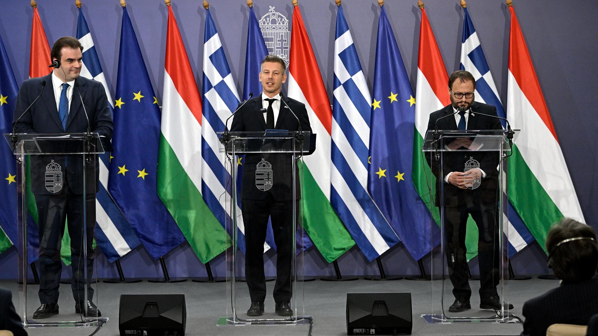 Magyar, Karman und Pierrakakis stehen bei einer Pressekonferenz nebeneinander an drei Rednerpulten. Im Hintergrund sind die Flaggen Ungarns, Griechenlands und der EU aufgestellt.