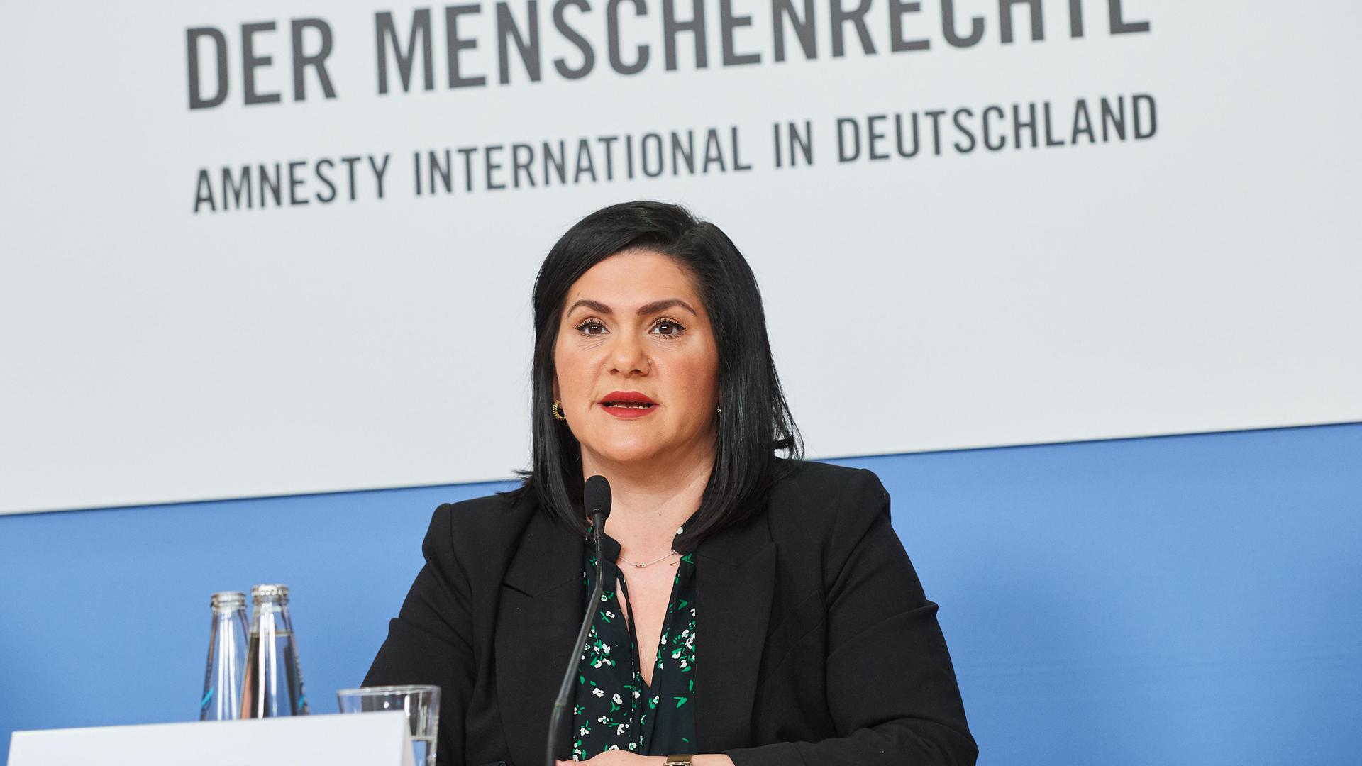 Mariam Claren sitzt bei einer Veranstaltung von Amnesty International Deutschland an einem Rednerpult. Sie hat schwarze, schulterlange Haare und trägt einen Blazer.