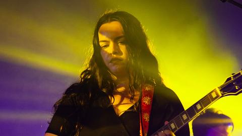 Eine junge Frau mit langen braunen Haaren steht in lila und gelbem Scheinwerferlicht und spielt ihre E-Gitarre.