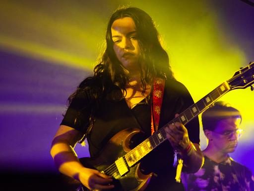 Eine junge Frau mit langen braunen Haaren steht in lila und gelbem Scheinwerferlicht und spielt ihre E-Gitarre.