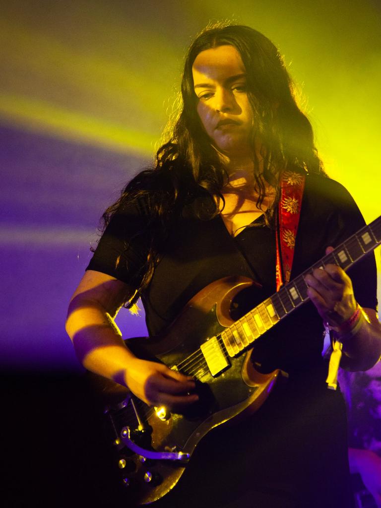 Eine junge Frau mit langen braunen Haaren steht in lila und gelbem Scheinwerferlicht und spielt ihre E-Gitarre.