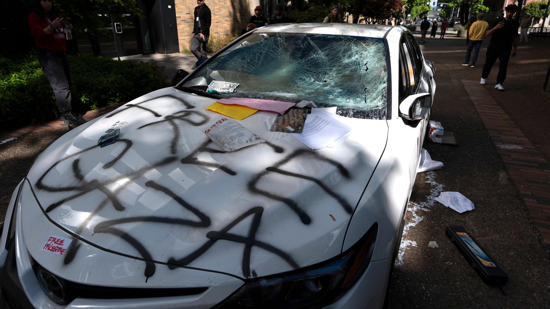 Die Scheiben des weißen PKW sind eingeschlagen. Auf der Kühlerhaube steht mit schwarzer Schrift gesprayt "Free Gaza". Dahinter mehrere Personen.