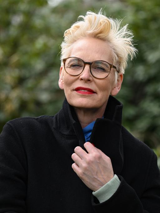 Bärbel Schäfer steht mit dunklem Mantel, dunlker Brille und knallrotem Lippenstift in einem Park und schaut in die Kamera.