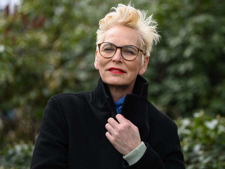 Bärbel Schäfer steht mit dunklem Mantel, dunlker Brille und knallrotem Lippenstift in einem Park und schaut in die Kamera.