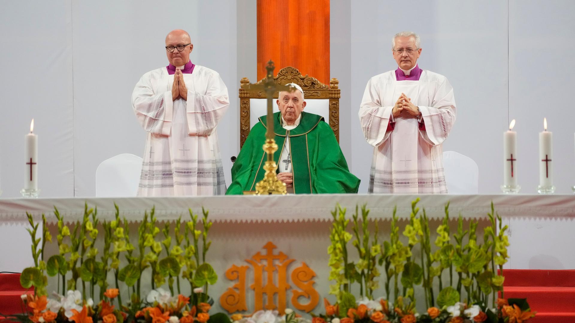 Der Papst sitzt im grünen Ornat an der Mitte des Altars vor einem Kreuz. An beiden Seiten jeweils ein Geistlicher in weiß, der die Hände zum Gebet gefaltet hat.