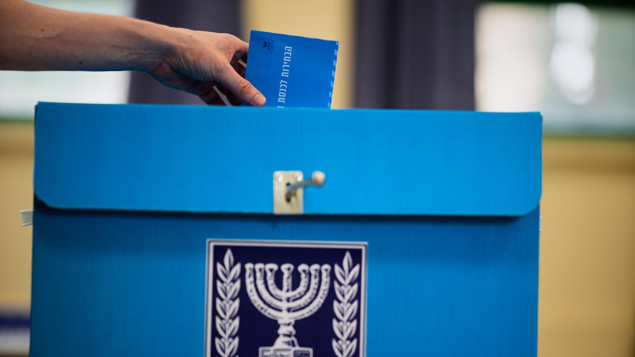 Eine Hand wirft einen blauen Umschlag ineiner große blaue Wahlurne. Auf der Vorderseite ist ein weißes Emblem mit Olivenzweigen zu sehen. Eine Hand wirft einen blauen Umschlag ineiner große blaue Wahlurne. Auf der Vorderseite ist ein weißes Emblem mit Olivenzweigen zu sehen.