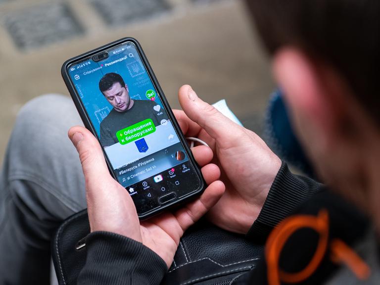 Ein ukrainischer Flüchtling hält ein Smartphone in der Hand und sieht sich das Video einer Rede von Wolodymr Selenskyi an.