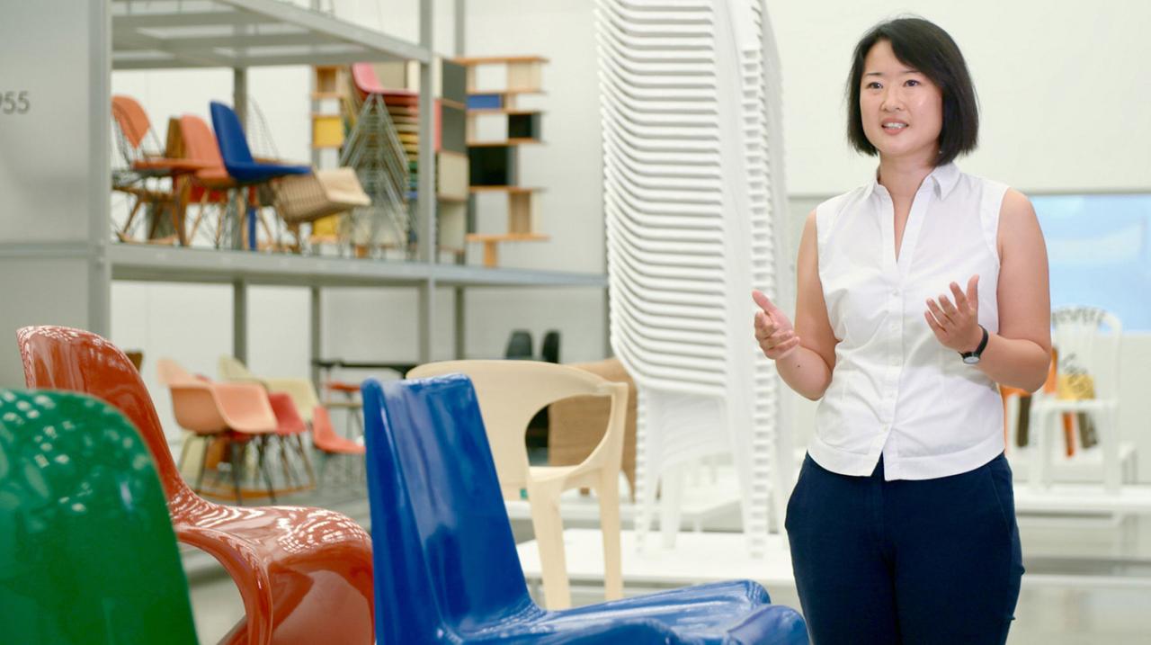 Eine asiatische Frau mit weißer Bluse und schwarzer Hose steht zur Kamera. Neben und hinter ihr bunte und gestapelte Plastikstühle. 