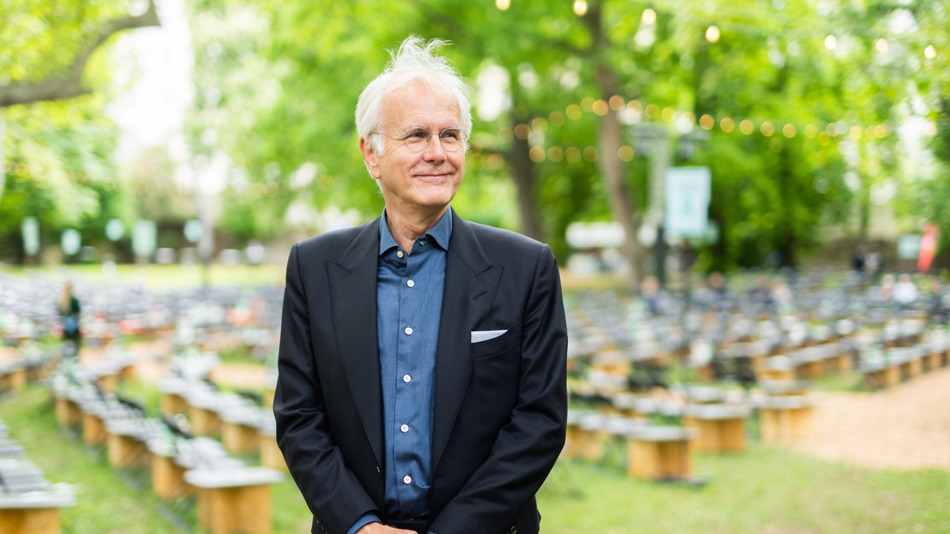 Harald Schmidt steht schmunzelnd und zur Seite schauend in einem Garten, im Hintergrund sind Sitzhocker zu sehen. Er trägt Brille, einen schwarzen Anzug und ein blaues Hemd.